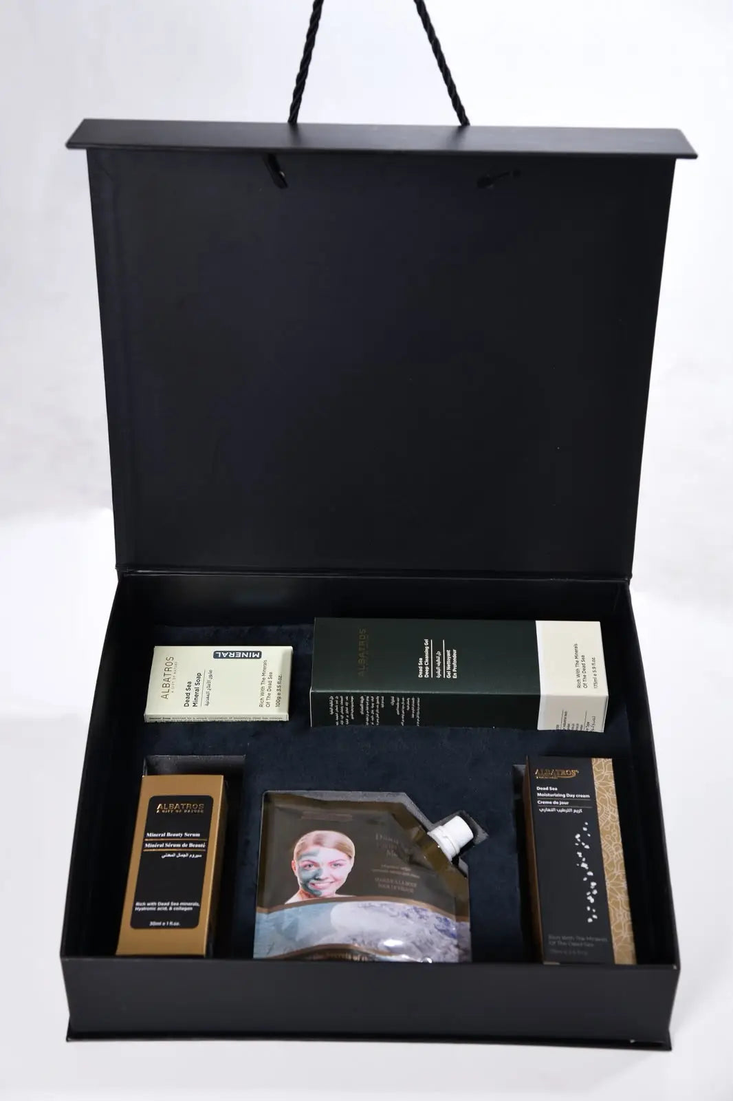 An open black gift box containing multiple skincare products, including a central sheet mask pouch, a dark green box, a beige box, and two slim brown boxes, photographed on a white background.