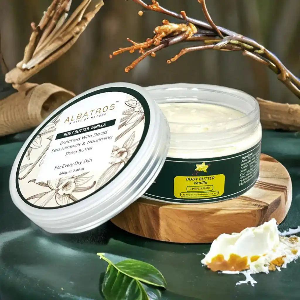 Albatros Body Butter Vanilla in a clear plastic jar with a dark green label, shown with its lid leaning beside it on a wooden stand. A dollop of the white body butter and a small leaf are placed in front, with dried branches in the background.